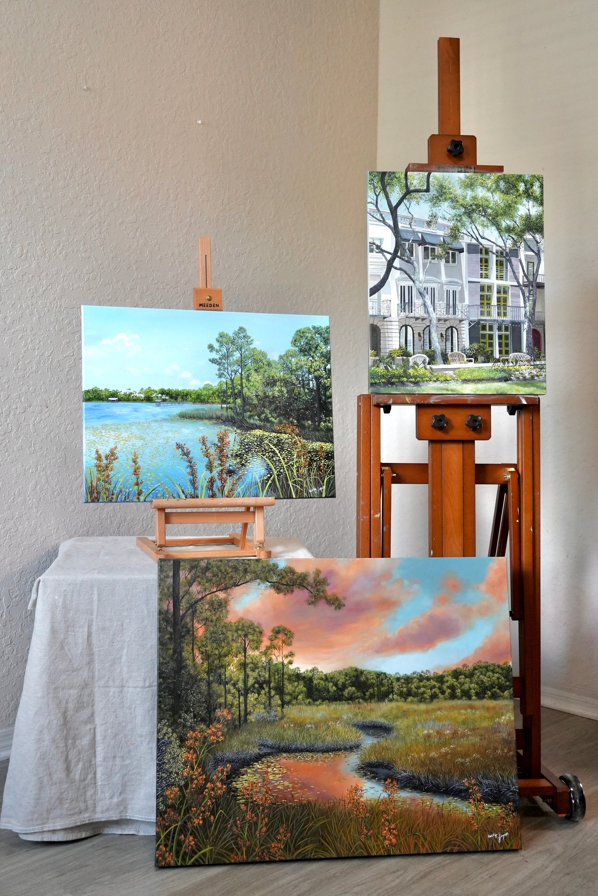 Three landscape paintings on easels against a wall.