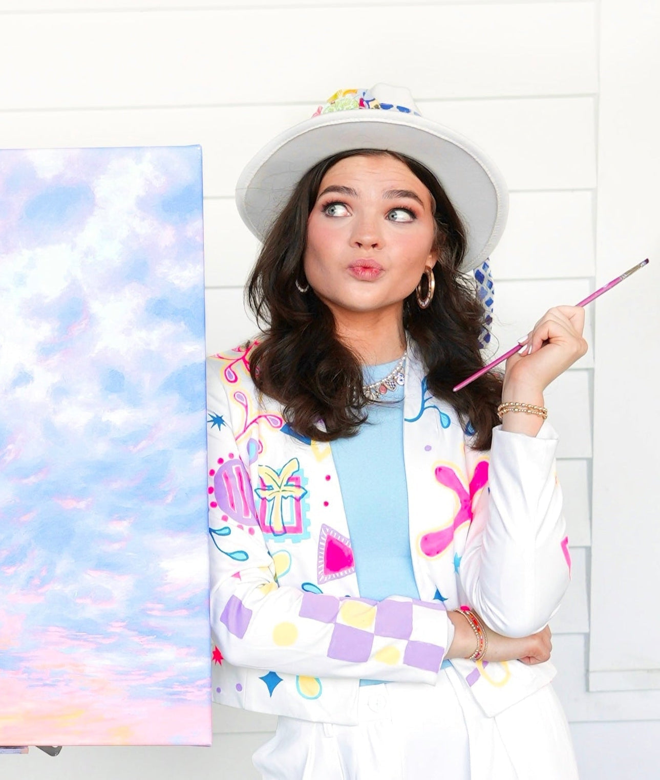 Woman wearing a colorful jacket and hat, holding a paintbrush with a painting of a sky and clouds in the background.