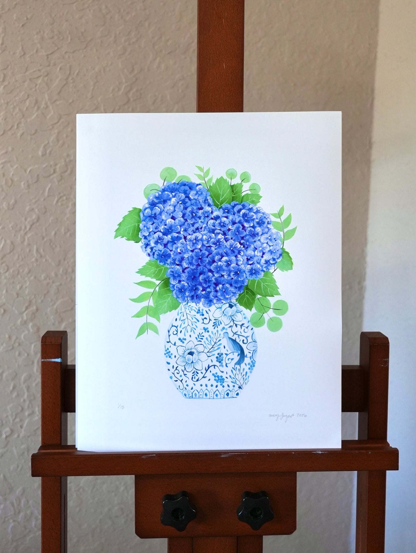 Art print of blue flowers in a vase on a wooden easel against a beige wall.