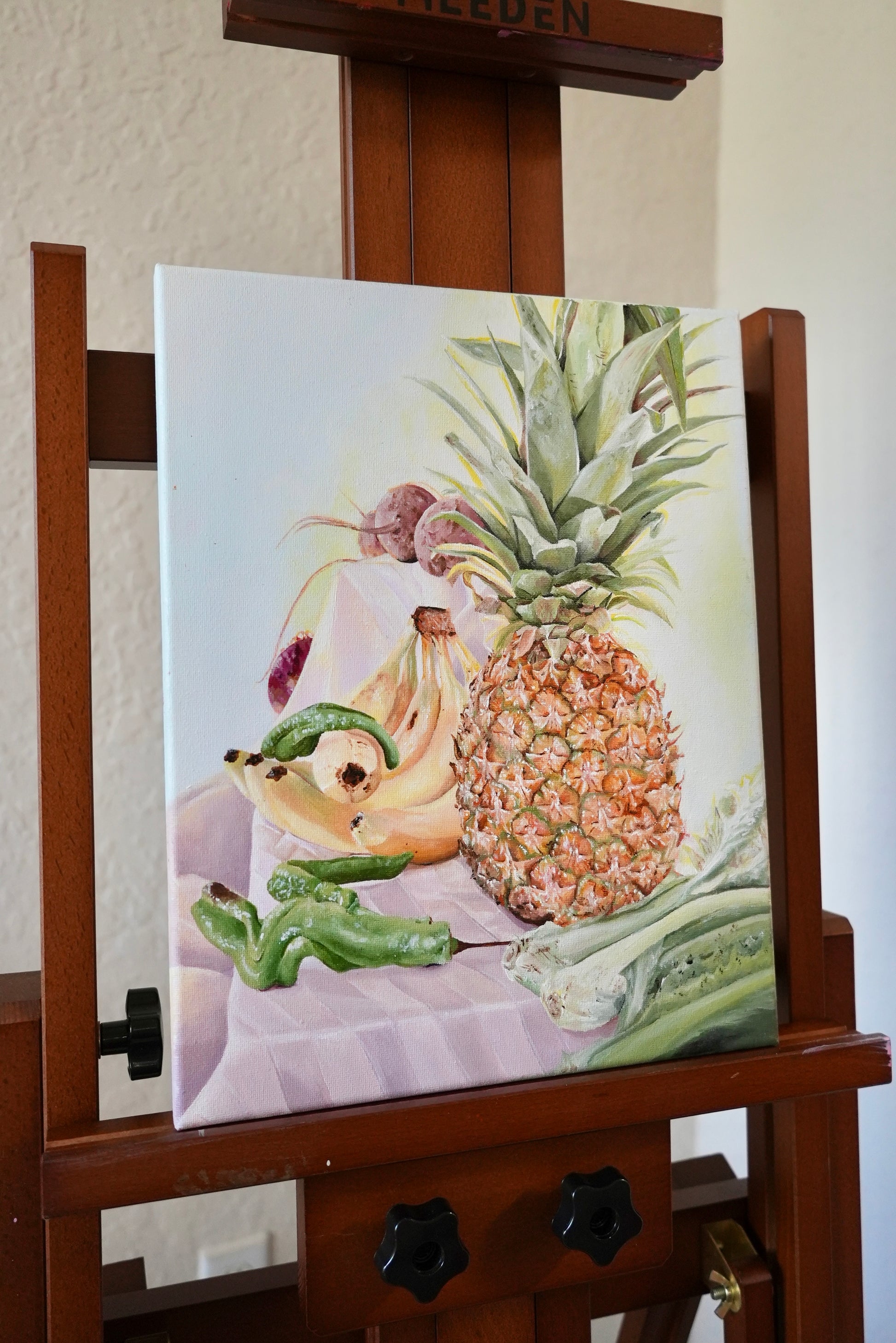 Painting of a pineapple and other fruits on an easel against a white wall.