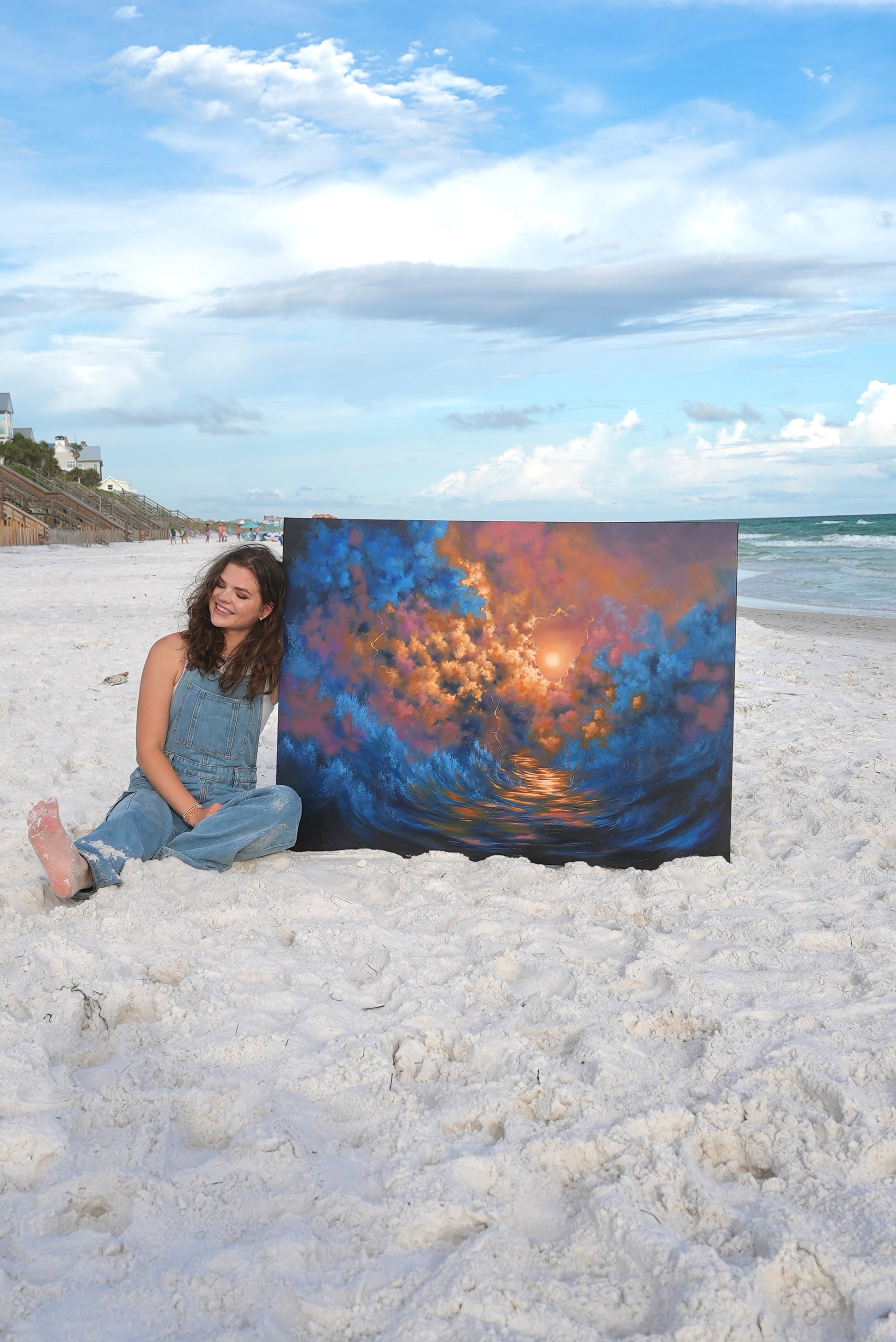 Avery sitting on a beach holding a large painting of a sunset over water.