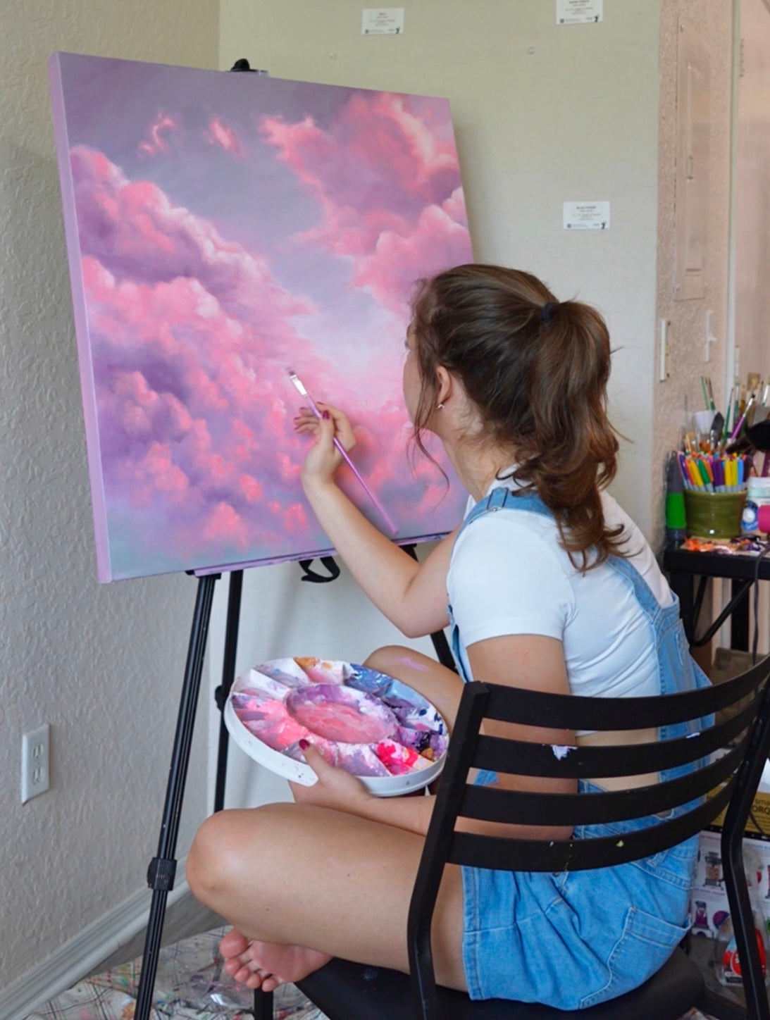 Avery painting a pink and purple cloud scene on an easel.
