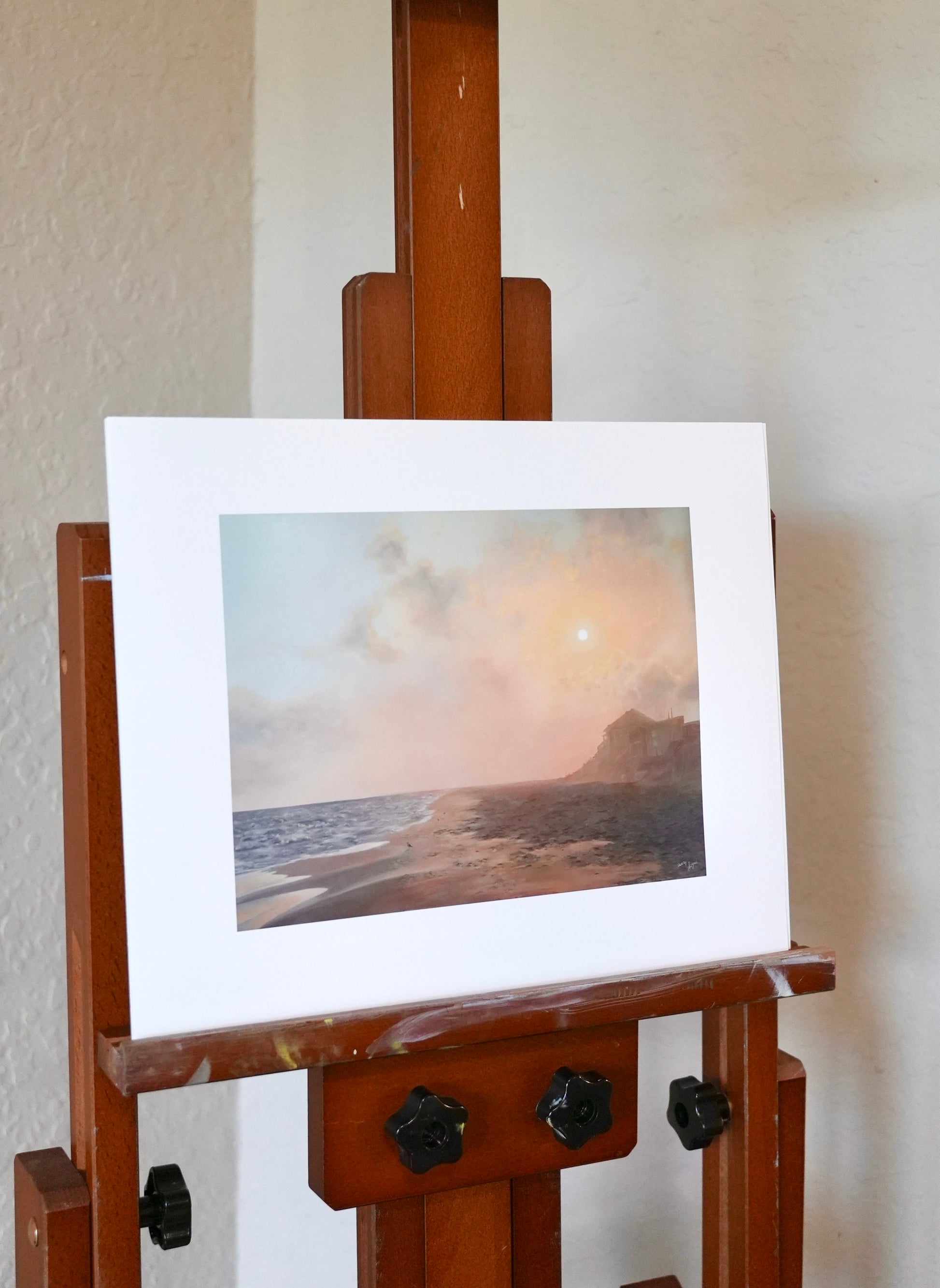 Wooden easel with an art print of a landscape on a white wall.