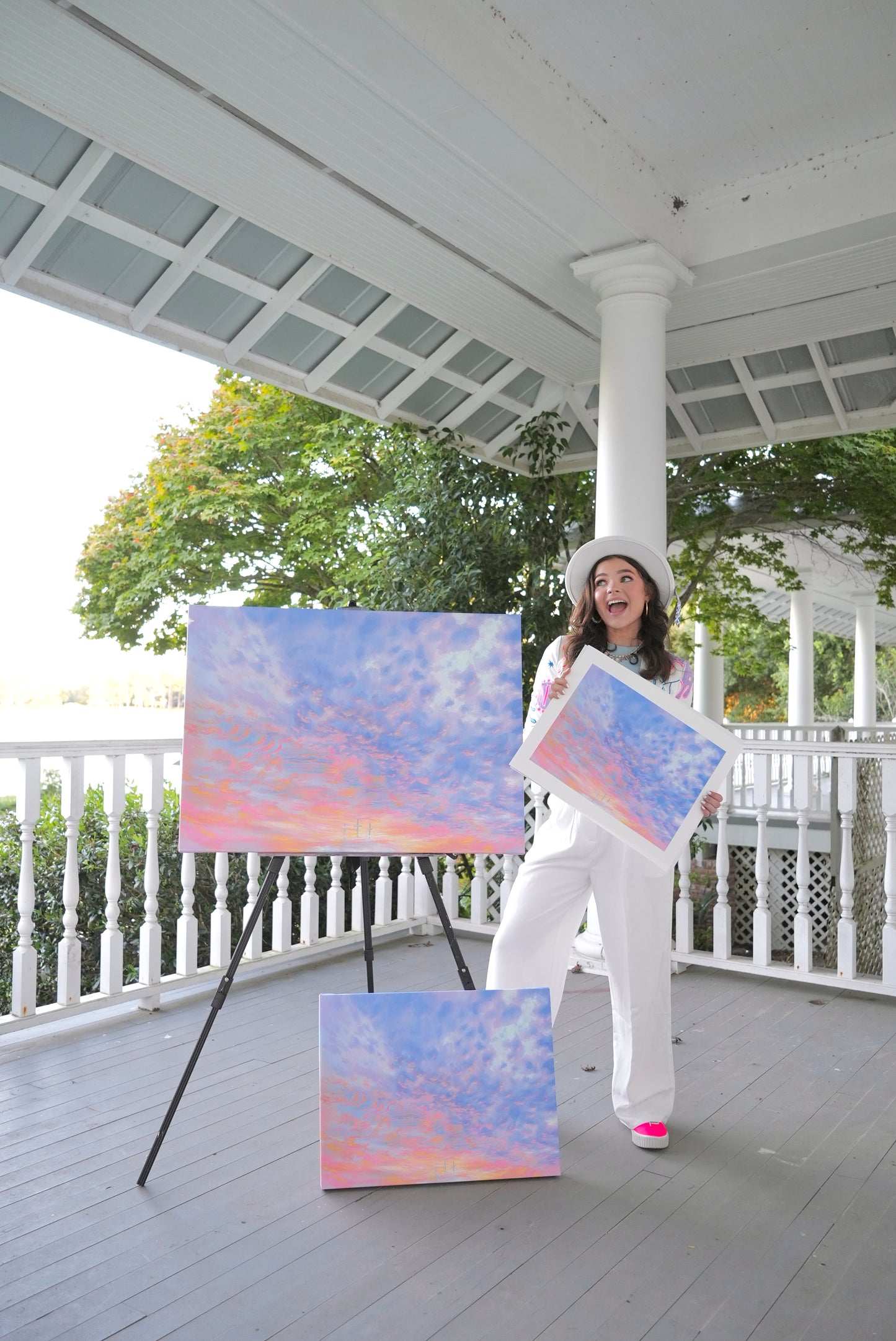 Avery holding a painting on a porch with another painting on an easel.