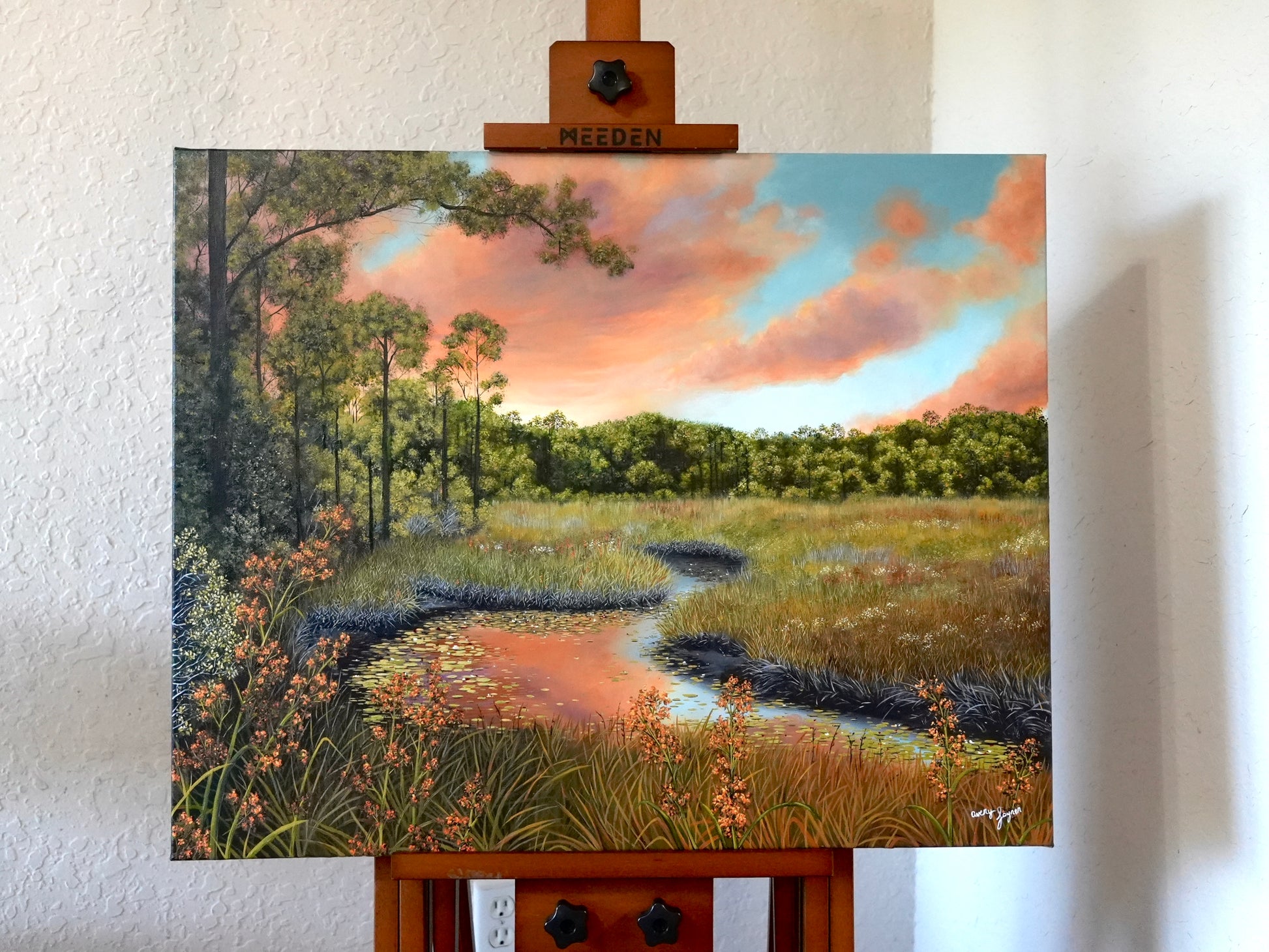 Painting of a landscape with trees, sky, and water on an easel.