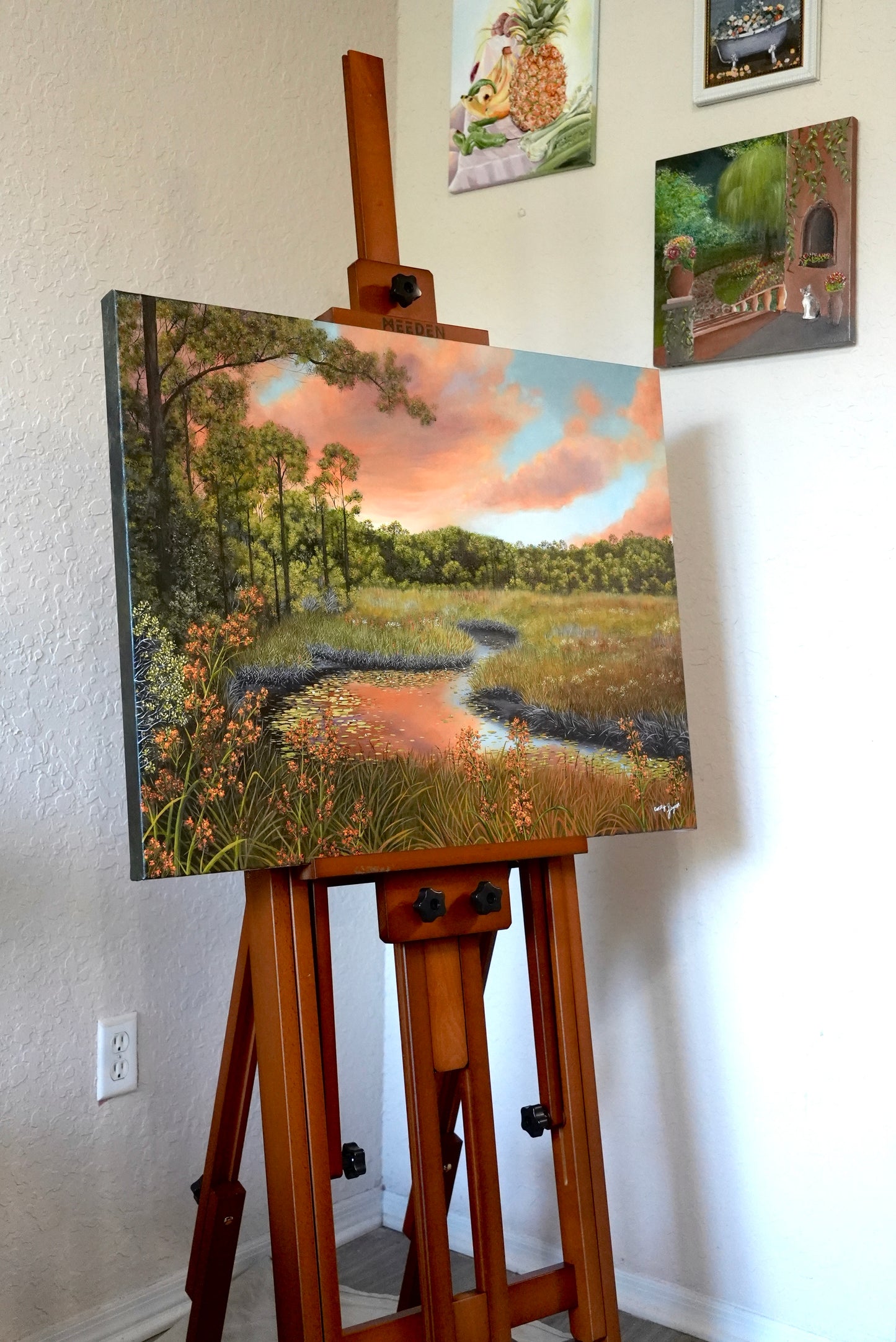 Painting of a landscape on an easel against a wall with other artworks.