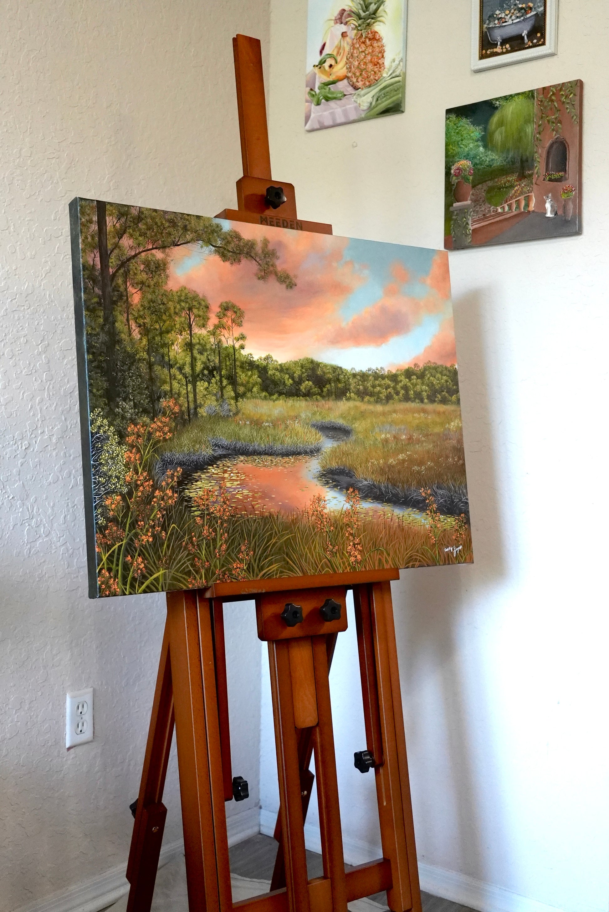Painting of a landscape on an easel against a wall with other artworks.