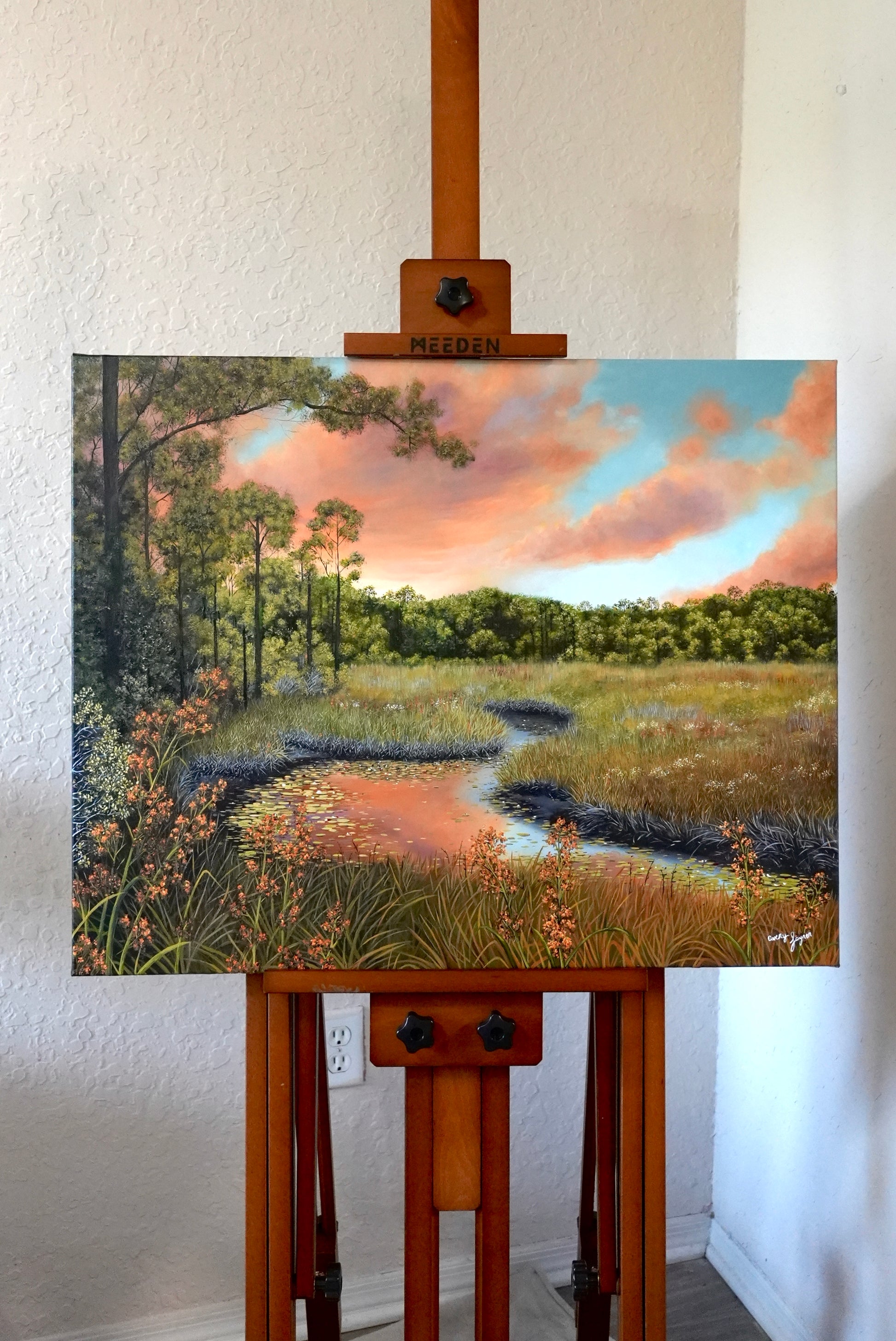 Painting of a landscape with trees and sky on an easel against a white wall.