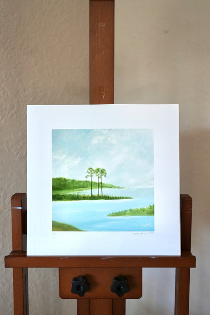 Framed artwork of a landscape with trees and water on a wooden easel.